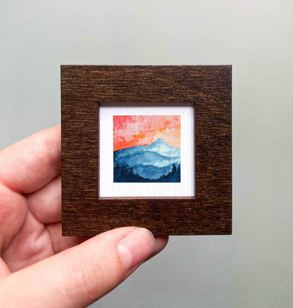 A hand is holding a small, square-shaped wooden frame containing a watercolor painting of a mountain landscape.