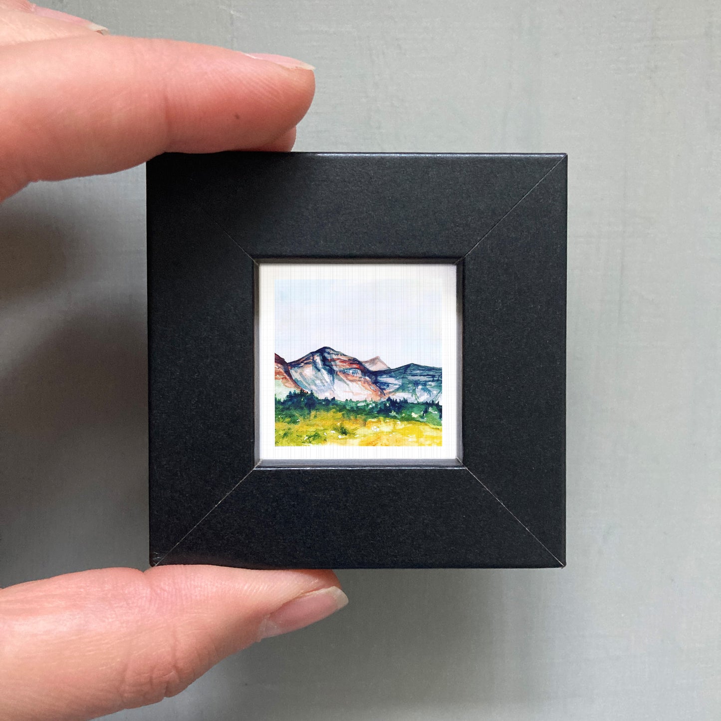 A person's hand holding a small black frame containing a watercolor painting of a mountain landscape.