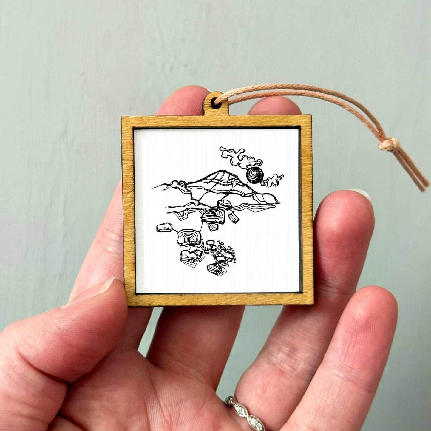 A hand holding a small wooden frame containing a black and white illustration of a mountain landscape.