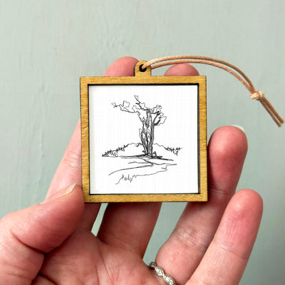 A hand holding a small wooden frame containing a black and white line drawing of a tree.