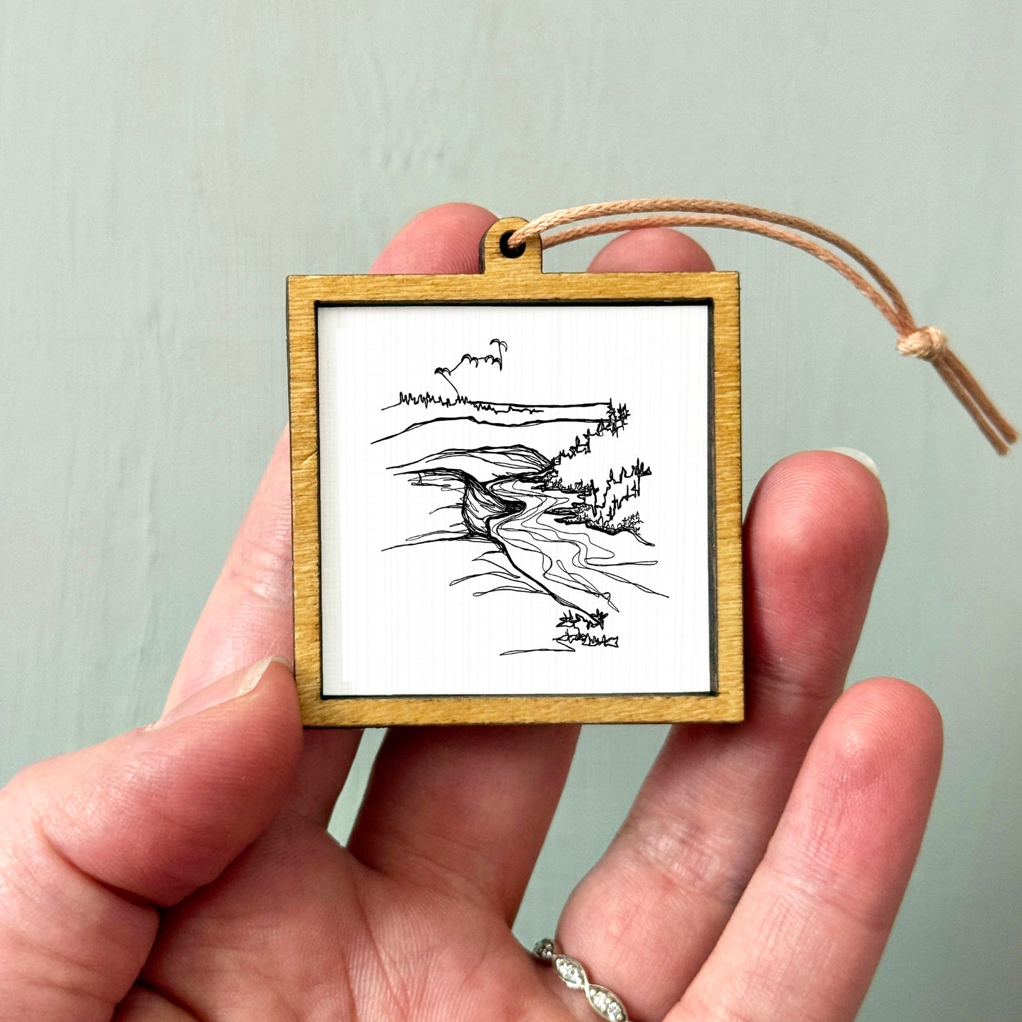 A hand holding a small wooden frame containing a black and white line drawing of a landscape.