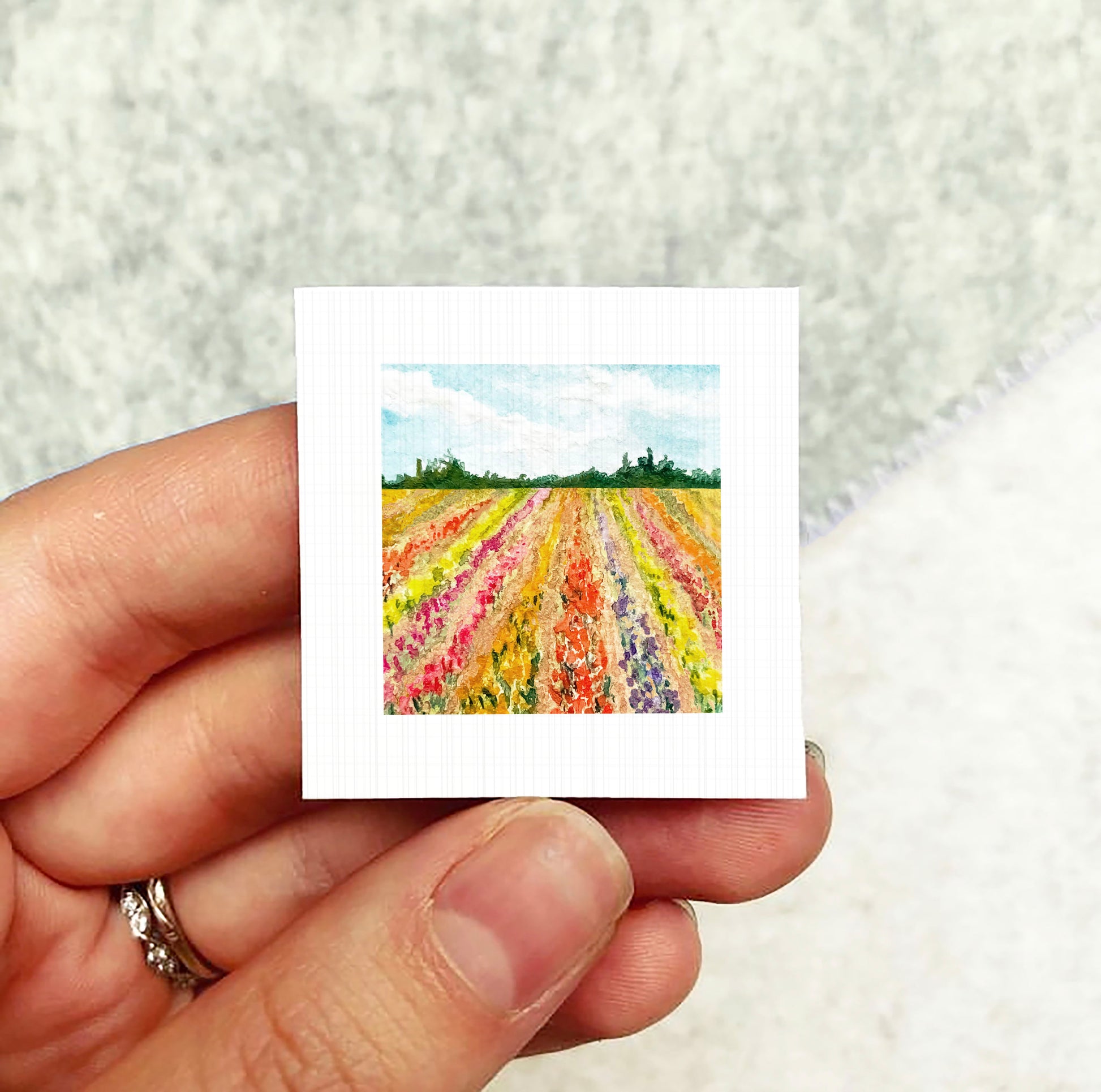 A hand holding a small square artwork depicting a colorful field of flowers.