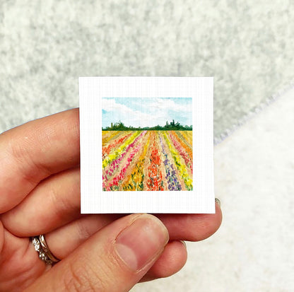 A hand holding a small square artwork depicting a colorful field of flowers.