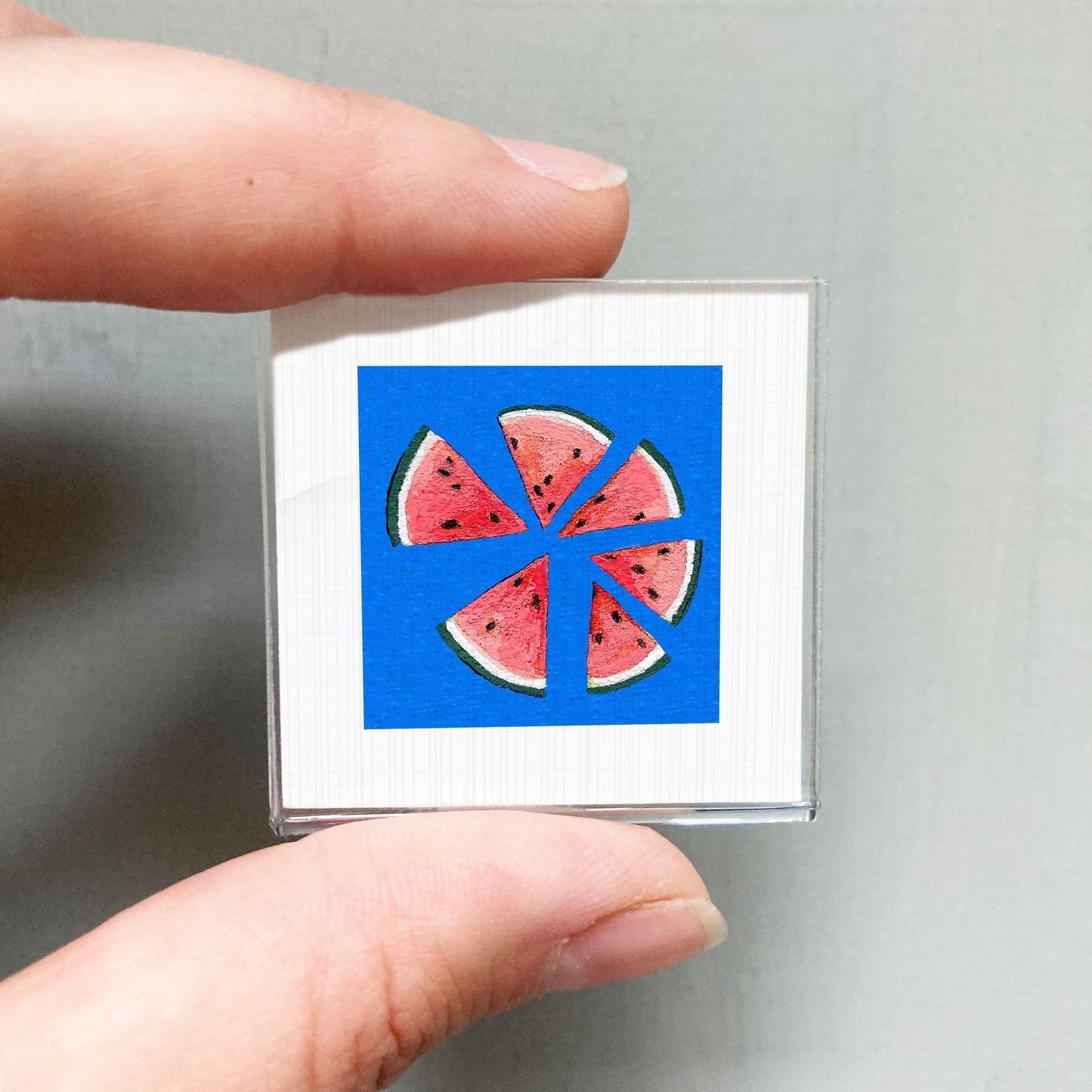A person's hand is holding a small square with a vibrant blue background and a watermelon slice design.
