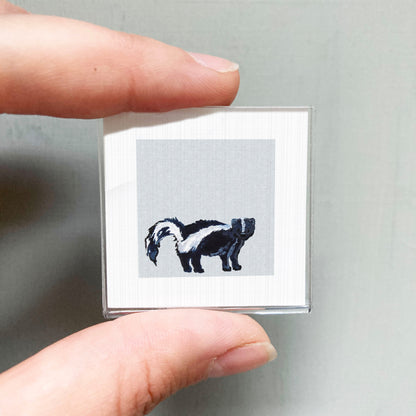 A person's hand is holding a small square picture featuring a black and white illustration of a skunk.