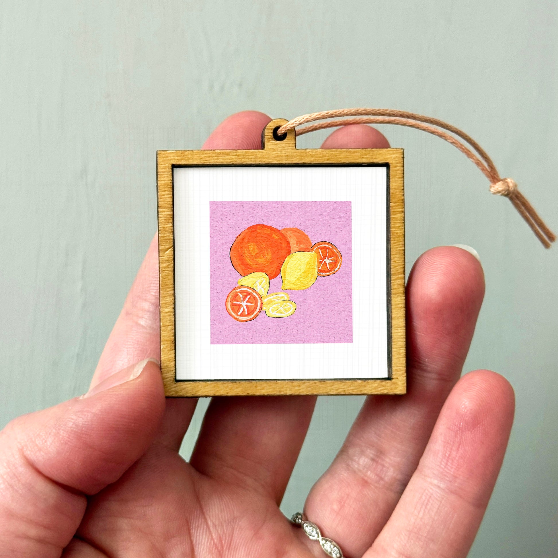 A hand holding a small wooden frame containing a framed illustration of various fruits, including oranges and lemons.
