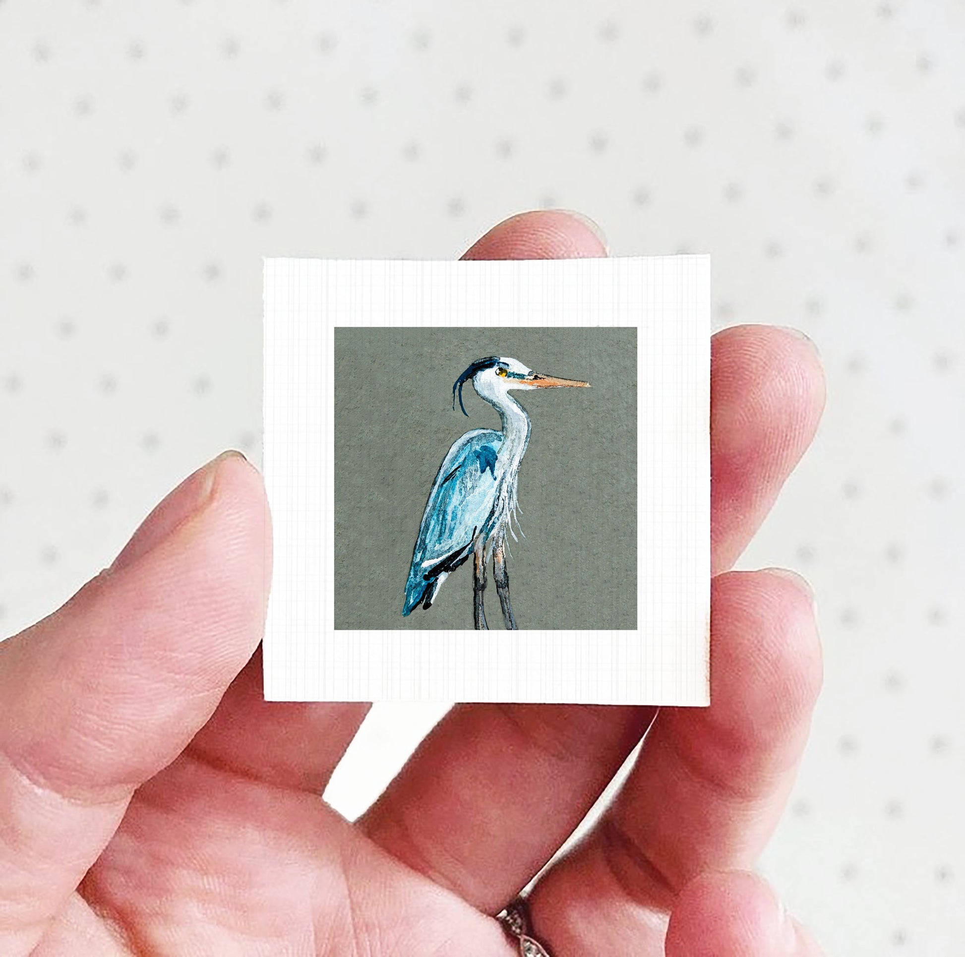 A hand holding a small square picture of a blue heron.