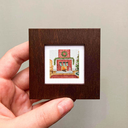 A hand is holding a small, square-shaped picture frame containing a painting of a fireplace with a wreath and stockings.