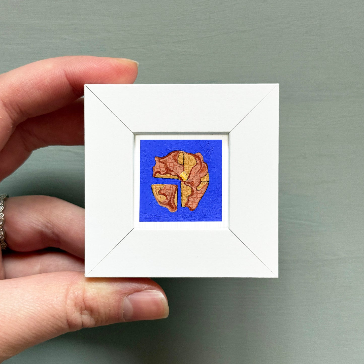 A hand is holding a small white square frame containing a vibrant blue and gold illustration of a lion's head.