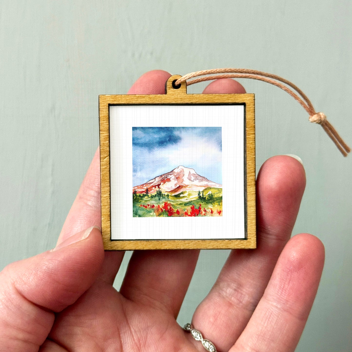 A hand holding a small wooden frame containing a watercolor painting of a mountain landscape.