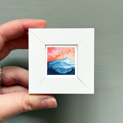 A hand is holding a small white square frame containing a watercolor painting of a mountain landscape with a sunset sky.