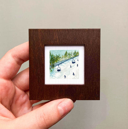 A hand is holding a small, square-shaped picture frame containing a miniature painting of a ski lift and people skiing.