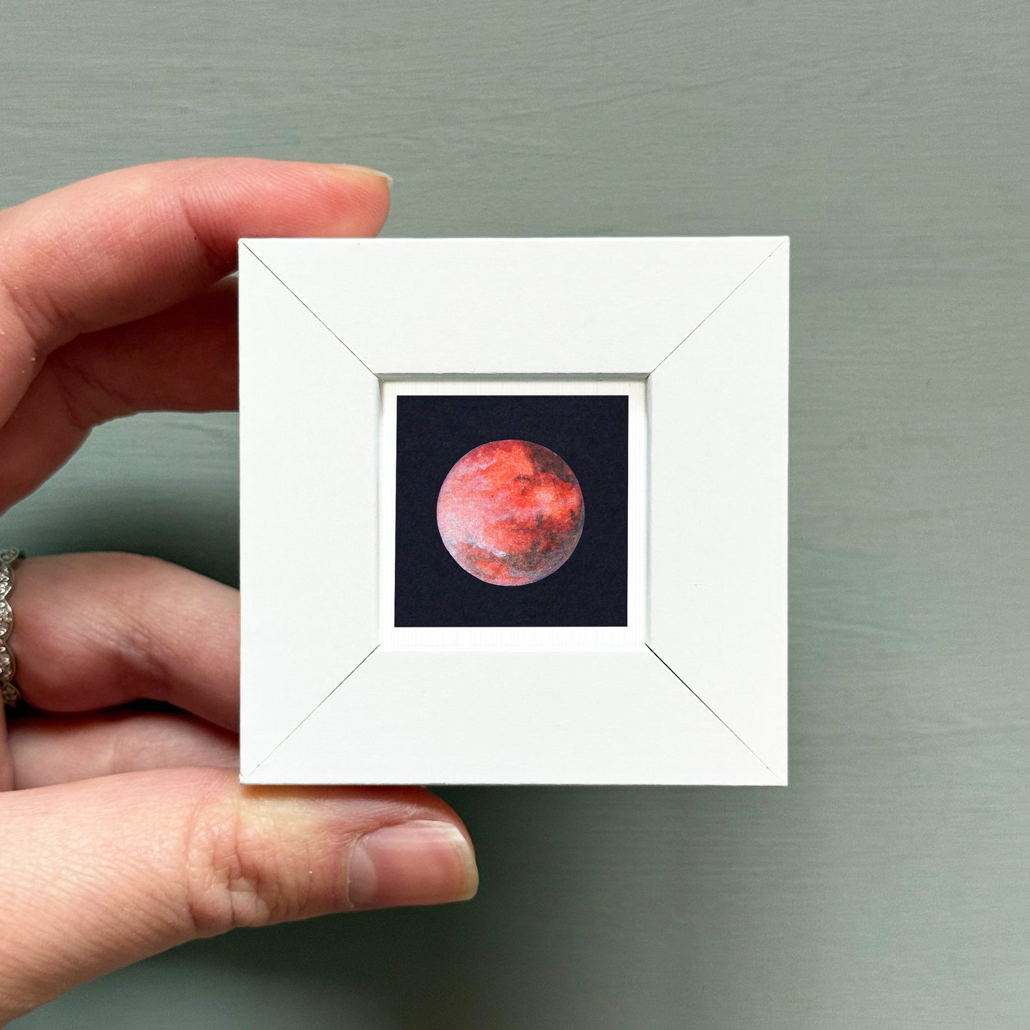 A hand is holding a small white square frame containing a framed image of a red planet, likely Mars, against a dark background.