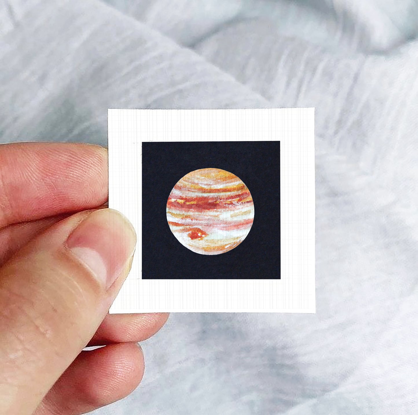 A hand is holding a small square-shaped paper with a picture of the planet Jupiter on it.
