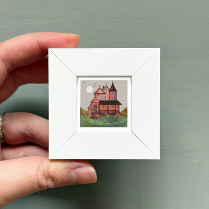A hand is holding a small white square frame containing a miniature painting of a house with a moon in the background.