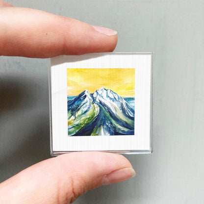 A person's hand is holding a small square picture of a mountain landscape with a yellow sky and green and blue colors.