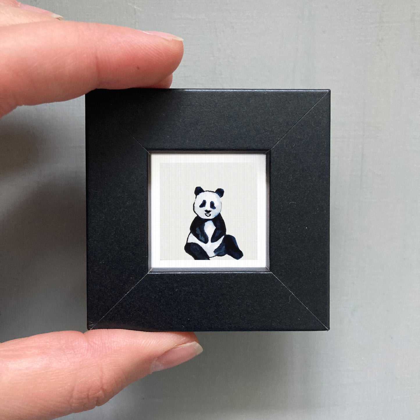 A person's hand is holding a small black square frame containing a picture of a panda bear.