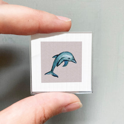 A person's hand is holding a small square picture of a dolphin.