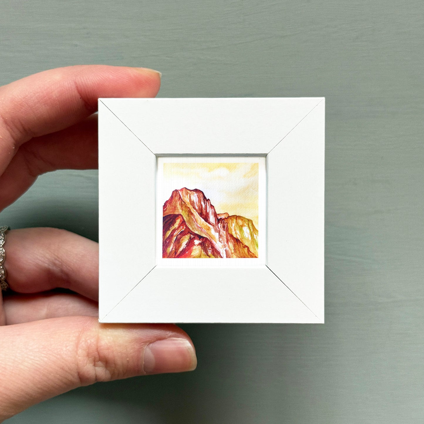 A hand is holding a small white square frame containing a painting of a mountain landscape.