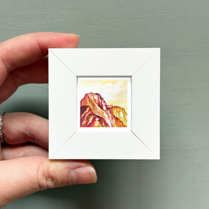 A hand is holding a small white square frame containing a painting of a mountain landscape.