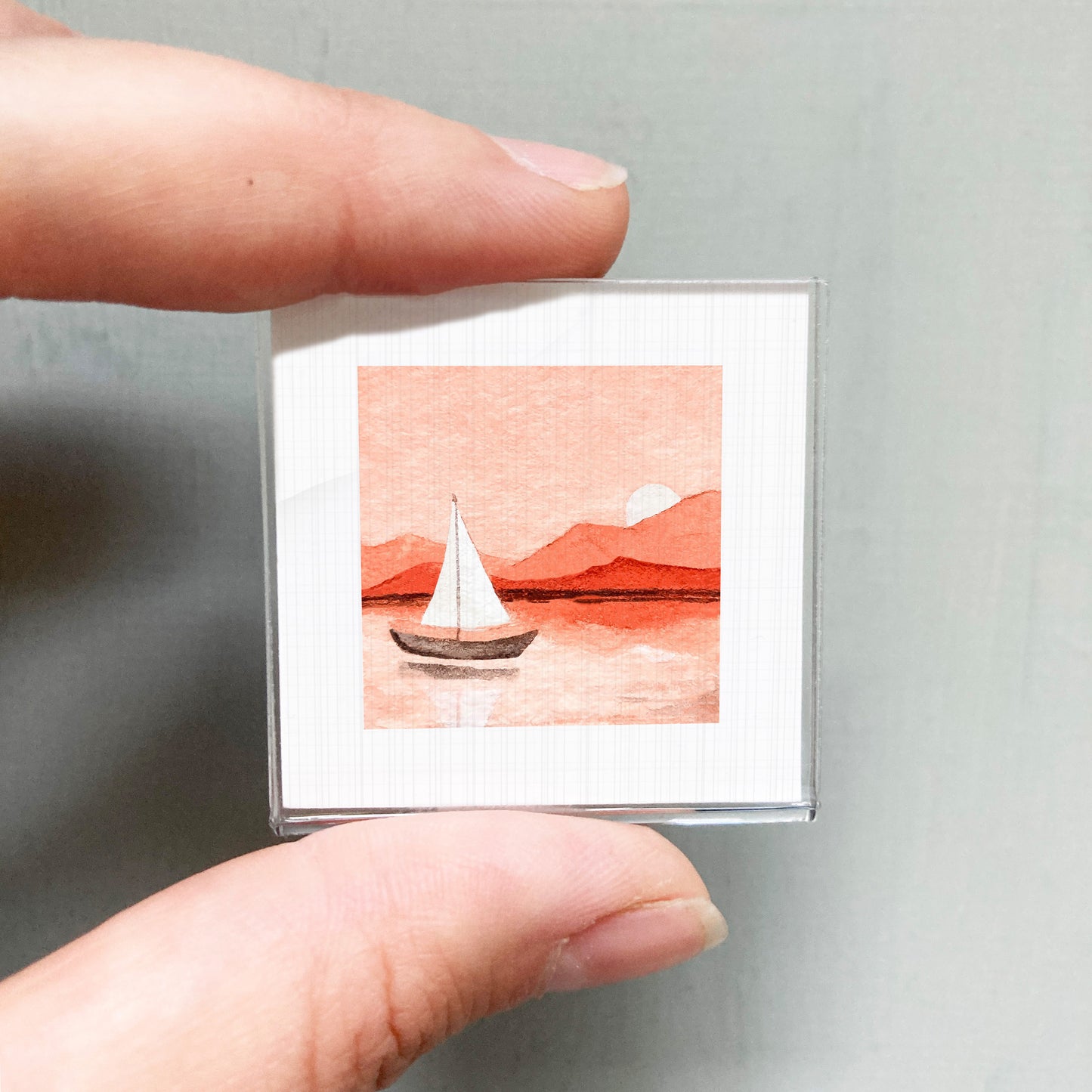 A hand is holding a small square picture depicting a sailboat on a body of water, with mountains in the background.