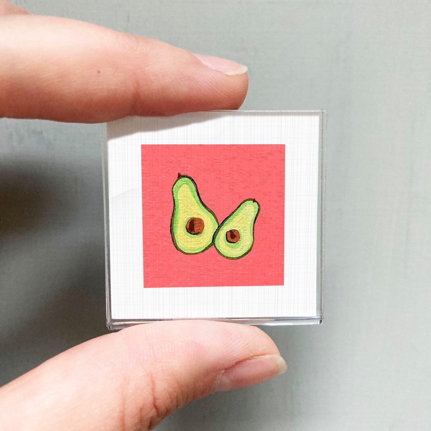 A person's hand holding a small square with a red background and two green avocados.