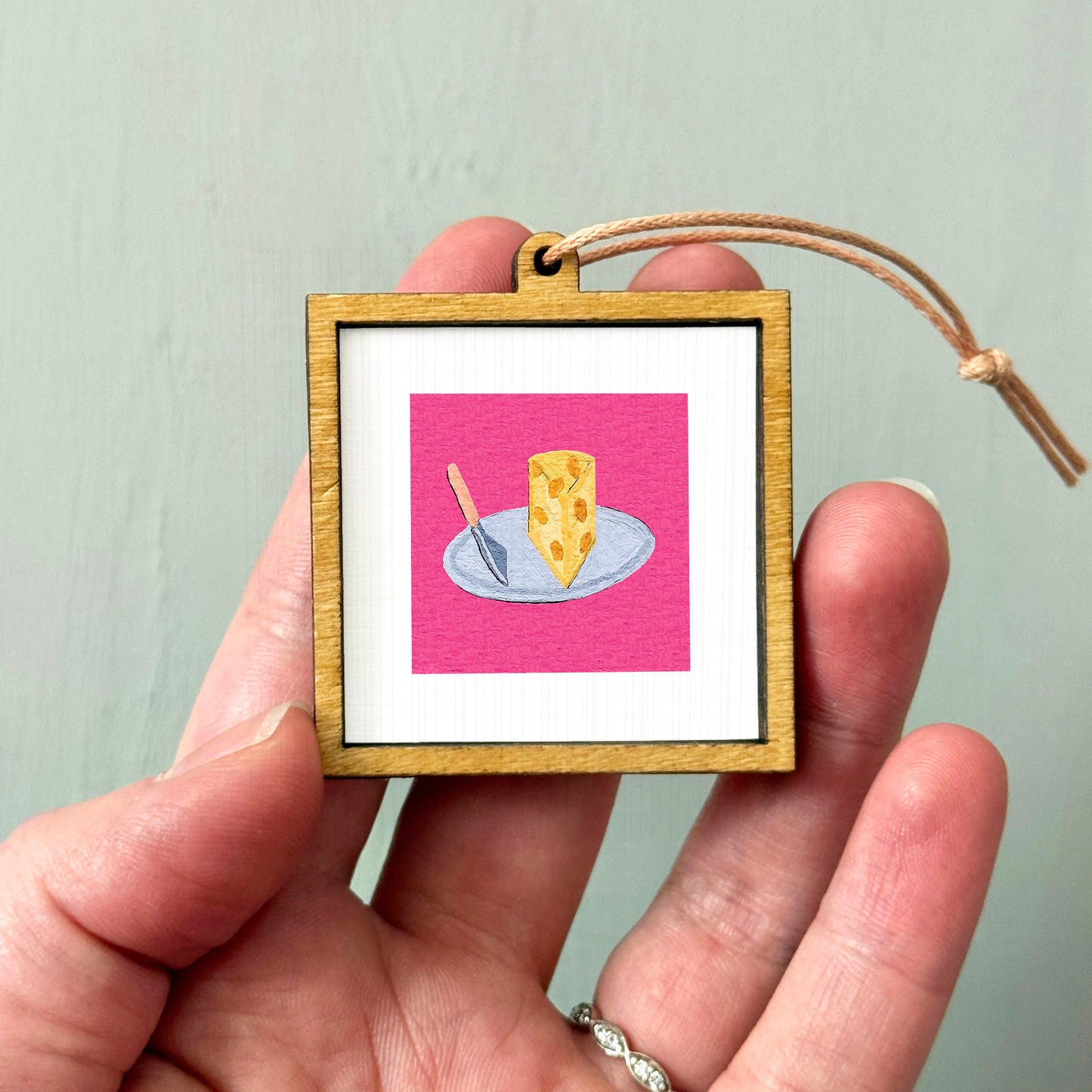 A person's hand holding a small wooden frame containing a picture of a piece of cheese on a plate.