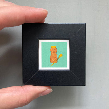 A person's hand is holding a small black picture frame containing a small illustration of a bear wearing a yellow hat.