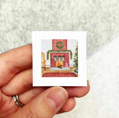 A hand holding a small square picture depicting a cozy fireplace scene with a Christmas tree and stockings.