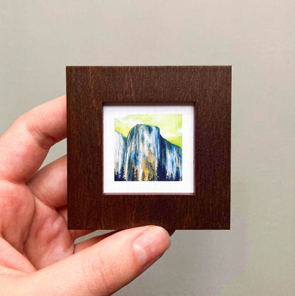 A hand is holding a small wooden frame containing a framed painting of a mountain landscape.