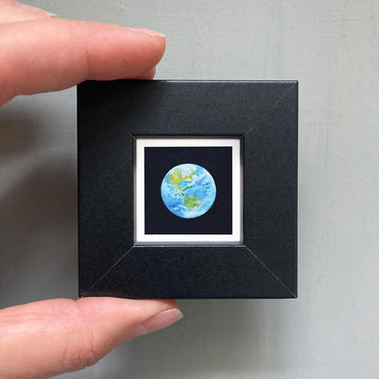 A person's hand holding a small black square frame containing a miniature representation of the Earth.