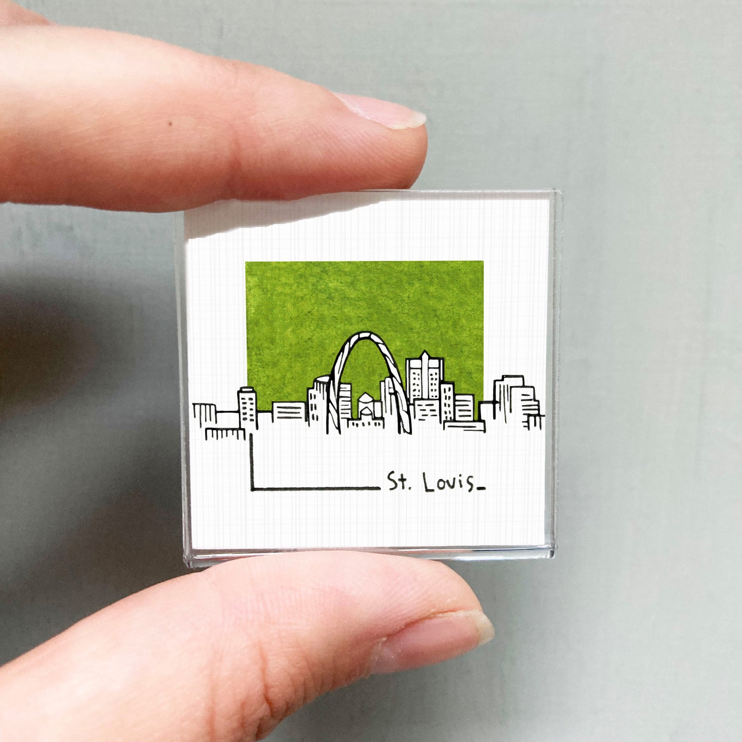 A hand holding a small square magnet with a drawing of St. Louis' skyline on it.