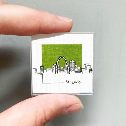 A hand holding a small square magnet with a drawing of St. Louis' skyline on it.