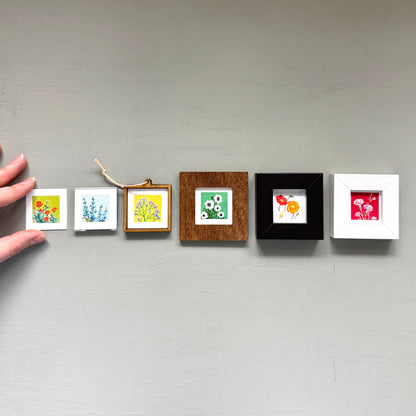 A hand is holding a row of five small framed artworks, each with a different color and style, against a gray wall.