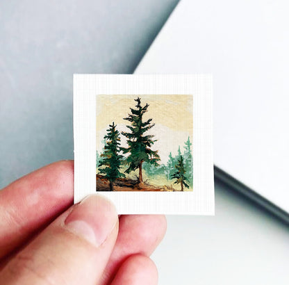 A hand is holding a small square painting of a forest scene with pine trees.