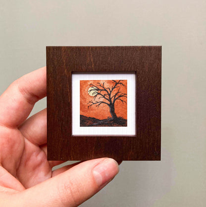 A hand is holding a small square frame containing a painting of a tree with a full moon in the background.