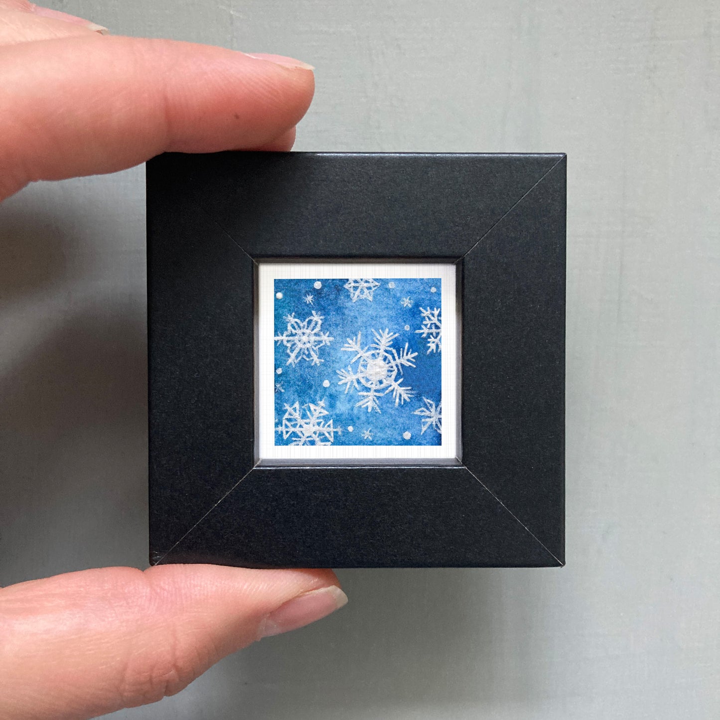 A hand is holding a small black square frame containing a picture of a blue background with white snowflakes.