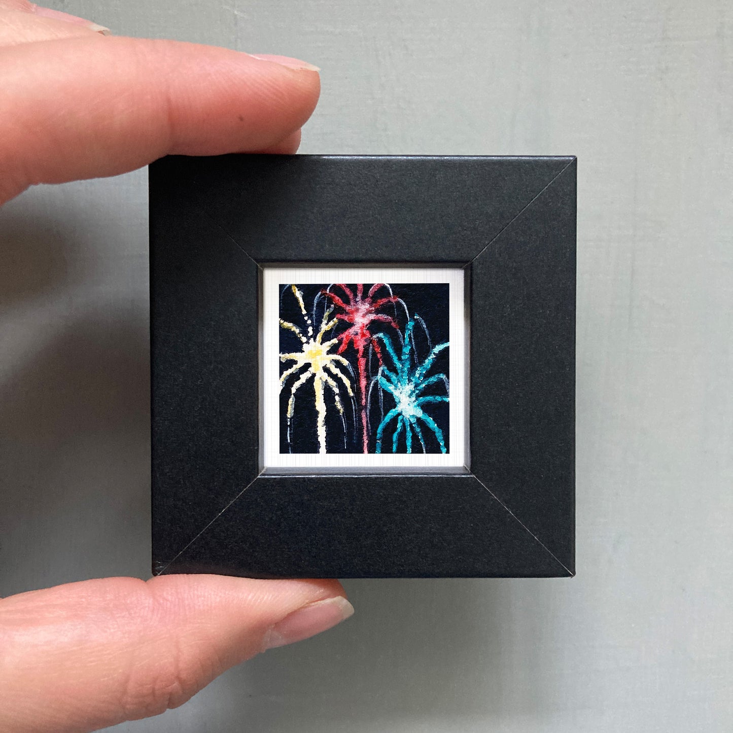 A hand is holding a small black square frame containing a vibrant, colorful fireworks display.