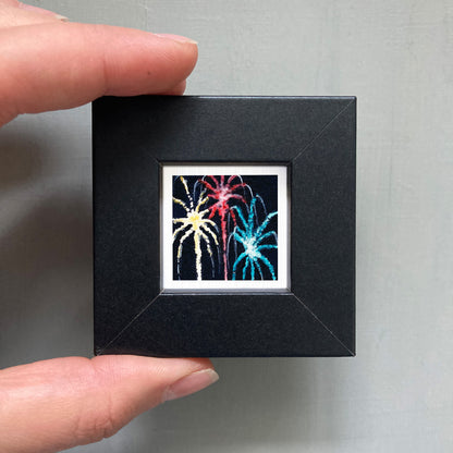 A hand is holding a small black square frame containing a vibrant, colorful fireworks display.