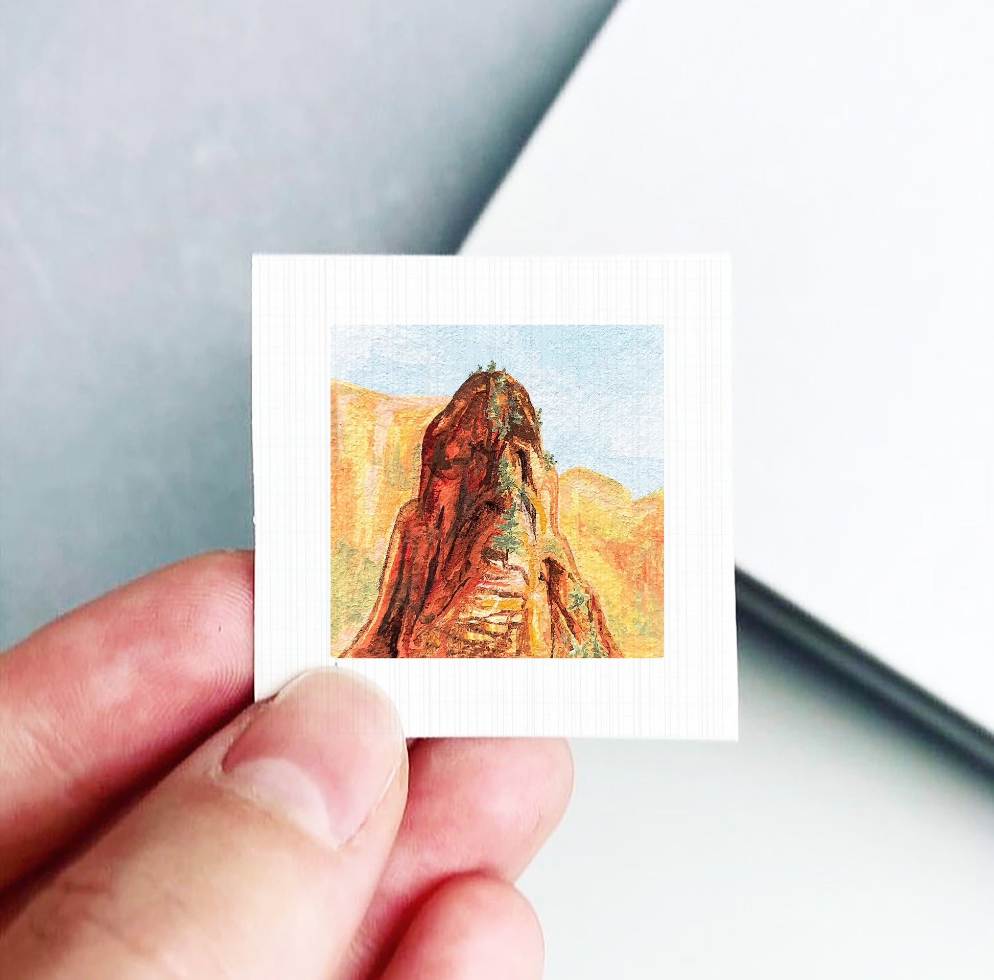 A hand holding a small square artwork depicting a mountain landscape.