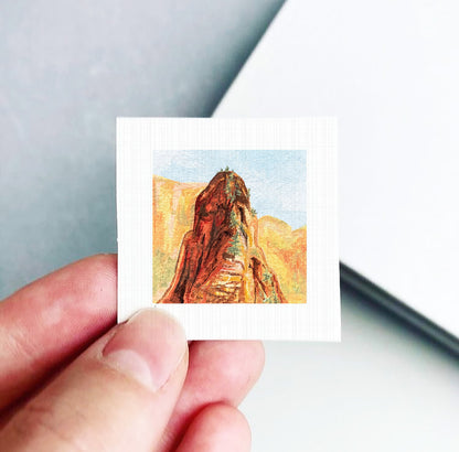 A hand holding a small square artwork depicting a mountain landscape.