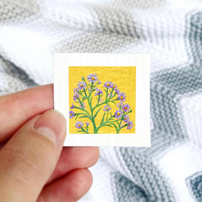 A hand is holding a small square card with a vibrant yellow background featuring a painting of purple flowers.