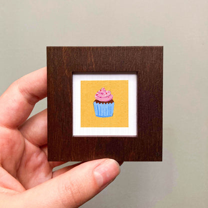 A hand is holding a small wooden frame containing a framed illustration of a cupcake with pink frosting.