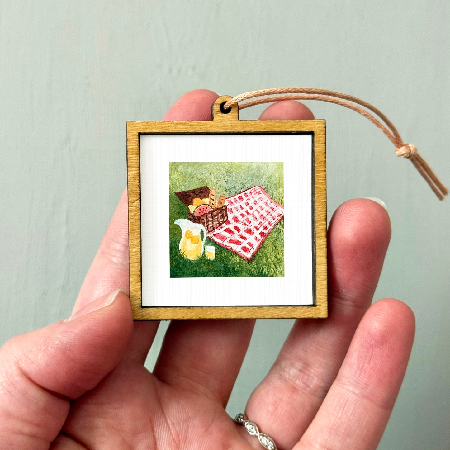 A hand holding a small wooden picture frame containing a painting of a picnic scene with a basket, blanket, and food items.