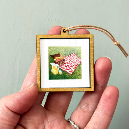 A hand holding a small wooden picture frame containing a painting of a picnic scene with a basket, blanket, and food items.