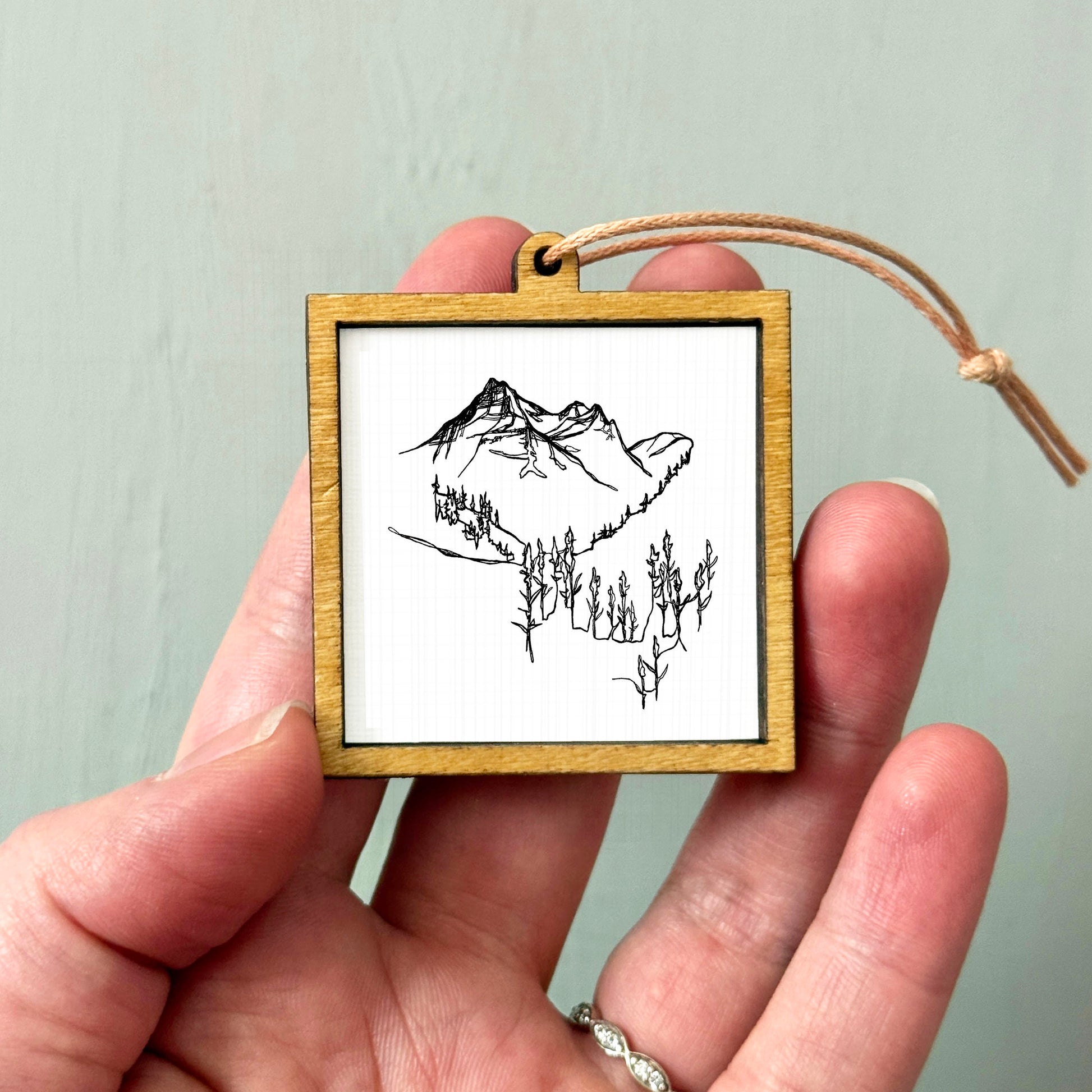 A hand holding a small wooden frame containing a black and white line drawing of a mountain landscape.