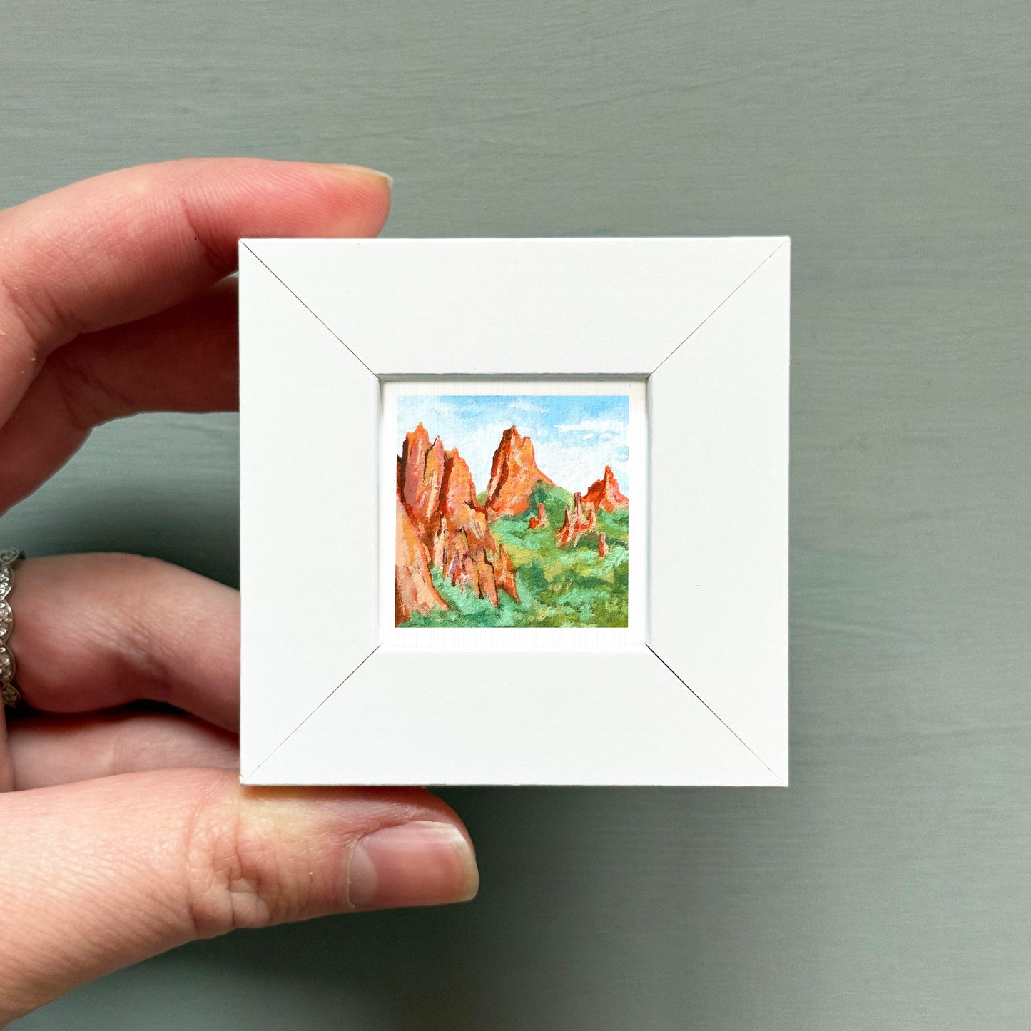 A hand is holding a small white square frame containing a painting of a mountain landscape.