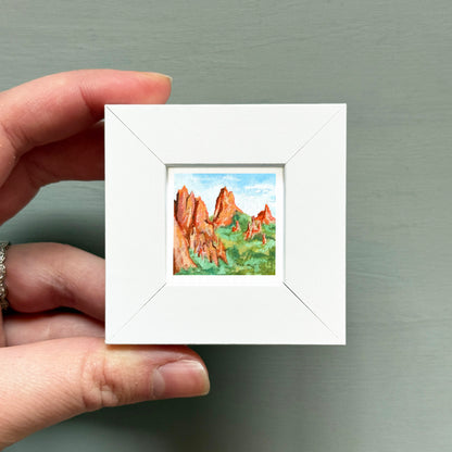 A hand is holding a small white square frame containing a painting of a mountain landscape.
