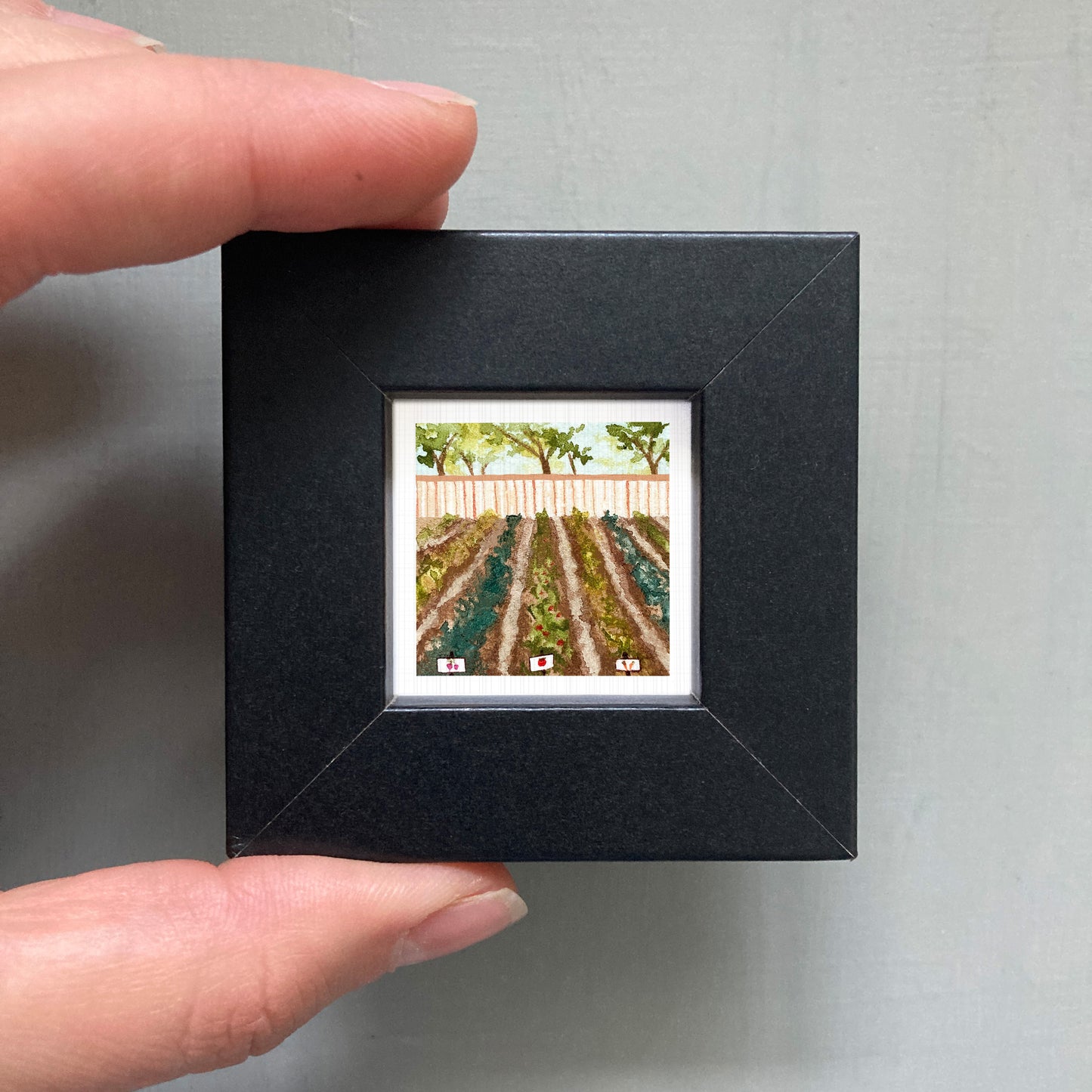 A person's hand holding a small black picture frame containing a painting of a field with trees.