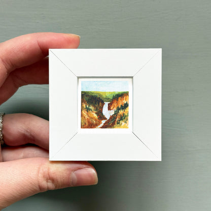 A hand is holding a small white square frame containing a painting of a waterfall in a forested area.